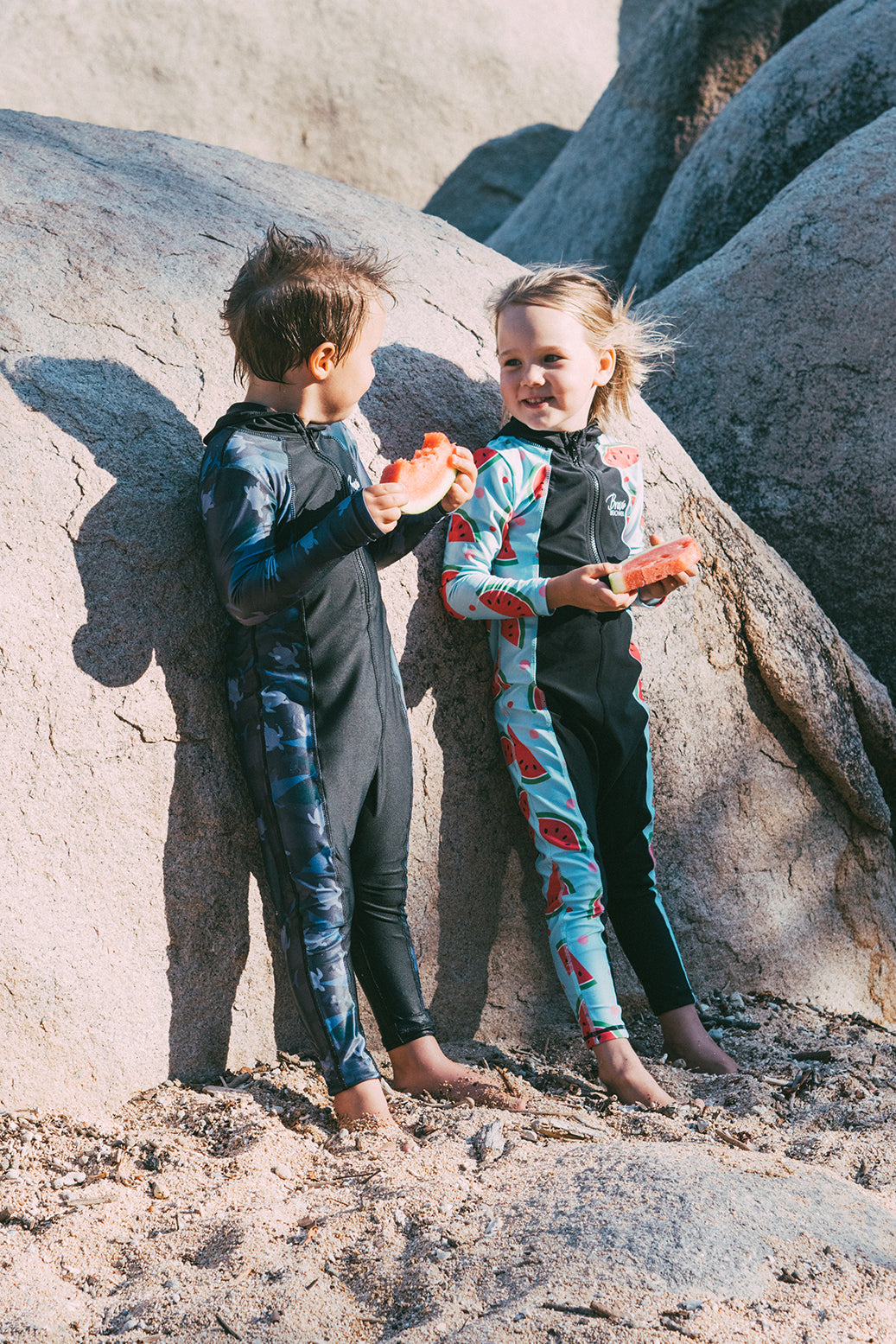 Boy and a girl in brave stingersuits eating watermelon and smiling at the beach